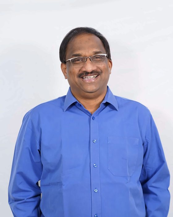 Prof. K. Nageshwar, smiling, in a blue shirt against a pale studio backdrop